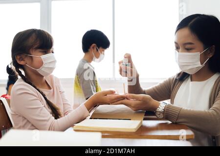 Junge Lehrerin, die ein Alkoholspray verwendet, um die Hände der Schüler im Klassenzimmer zu desinfizieren. Asiatische Frau in Gesichtsmaske Reinigung Pupillen Hände mit Hand Desinfektionsmittel. Schule nach Quarantäne und Sperrung wieder öffnen. Stockfoto