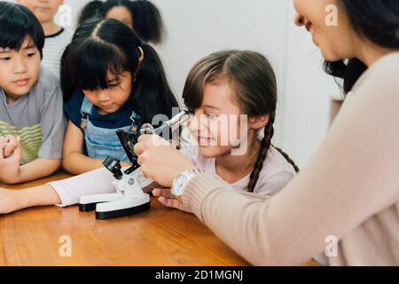 Junge Frau zeigt multiethnische Kinder Mikroskop in der Wissenschaft Klasse, Lernen, Entdeckung, Neugier. Asiatische Schule Lehrer hilft Schülern Mikroskop verwenden Stockfoto