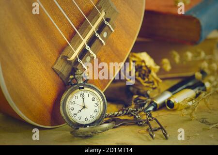 Eine antike Taschenuhr lehnte sich an eine Ukulele und ein altes Buch mit Vintage-Karte und Messingstift auf Holztisch platziert. Nahaufnahme und Kopie Platz für Text. Stockfoto