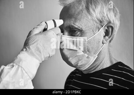 Nahaufnahme eines zugeschnittenen Schnappschusses der Hand des Arztes im Handschuh und eines PSA-Anzugs zur Messung des alten Männerfiebers mit Infrarot-Thermometer, Kopf des Patienten in Maske, schwarz-weiß. COVID-19 Konzept Stockfoto