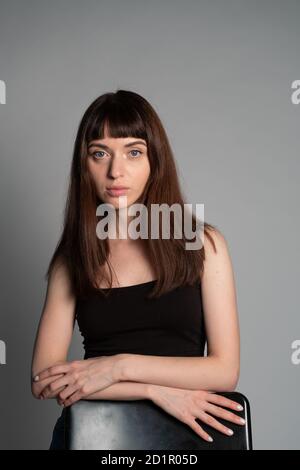 Studioportrait einer hübschen Brünette in einer schwarzen Weste, hinter dem Stuhl stehend, die Hände gekreuzt, ernsthaft auf die Kamera schauend, gegen eine Ebene Stockfoto