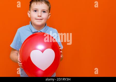 Nahaufnahme Valentinstag. Glücklich kaukasischen fröhlichen jungen hält einen großen roten Ballon.Kid mit einem weißen Herz auf Ballon gezeichnet.Orange Studio Hintergrund. Stockfoto