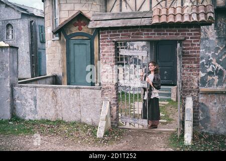 Junge Brünette Frau steht am Tor einer alten Kirche. Retro Kleidung an Halloween Stockfoto