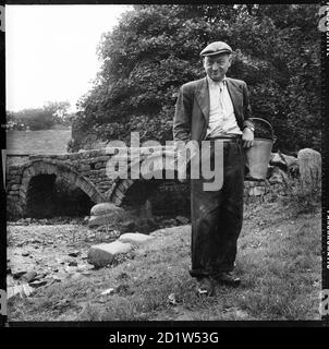 Ein informelles Porträt eines Mannes, der traditionelle Clogs trägt, einen Eimer hält, der vor der Packhorse Bridge in Wycoller, Trawden Forest, Pendle, Lancashire, Großbritannien, steht. Stockfoto