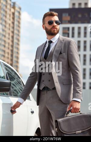 kaukasischer Geschäftsmann in formalwear, der die Tür seines repräsentativen Autos öffnet, bärtiger Mann in Sonnenbrille nach der Arbeit, Bürotasche in den Händen halten Stockfoto