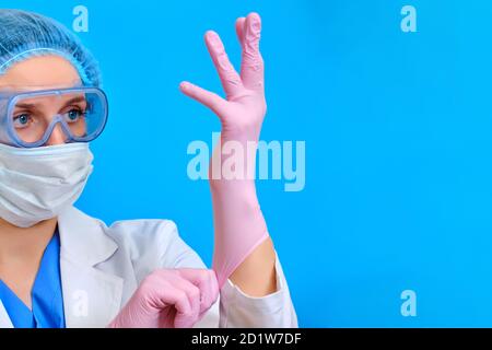 Der Arzt zieht Schutzhandschuhe an, um mit Patienten mit Coronavirus, blauer Hintergrund, zu arbeiten. Krankenschwester in einem weißen Mantel und schützende medizinische Ausrüstung Stockfoto