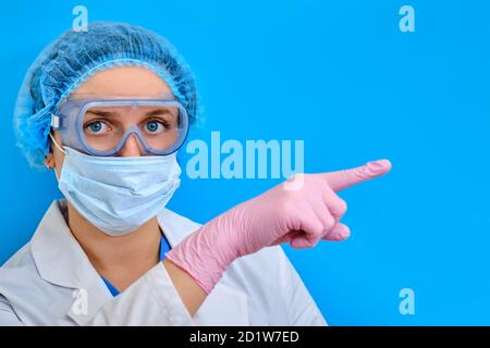 Der Arzt in medizinischen antiviralen Brillen zeigt einen Finger zur Seite, kopieren Sie Platz für Text auf einem blauen Hintergrund. Nahaufnahme der Krankenschwester in Schutzmaske und Stockfoto