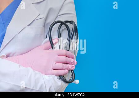 Eine Krankenschwester in schützender medizinischer Kleidung hält ein Stethoskop, blauer Hintergrund. Ein Arzt in einem weißen Mantel und rosa Handschuhe mit einem Stethoskop in den Händen, Stockfoto
