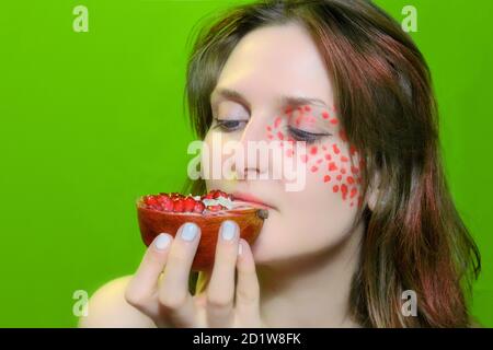 Weiblicher Gesichtsdruck von Make-up. Mädchen mit Obst und roten Make-up. Die Frau und die Hälfte des Granatapfels auf grünem Hintergrund. Stockfoto