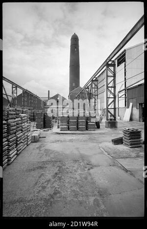 Allgemeine Ansicht des Elswick Shot Tower, abgerissen 1968-69, verwendet für die Erstellung von Bleiaufnahmen, mit einem Hof mit Paletten im Vordergrund Schusstürme wurden verwendet, um sphärische Aufnahmen von Blei zu erstellen. Dies wurde getan, indem man geschmolzenes Blei durch einen Seicve hinunter einen großen Schacht oder Turm in Wasser fallen ließ. Das Metall kühlt ab, wenn es fällt und dreht sich aufgrund der Oberflächenspannung kugelförmig. Dieser Schussturm war Teil der Elswick Bleiwerke, und der Turm wurde abgerissen 1968-69. , um 1955 - um 1969. Stockfoto