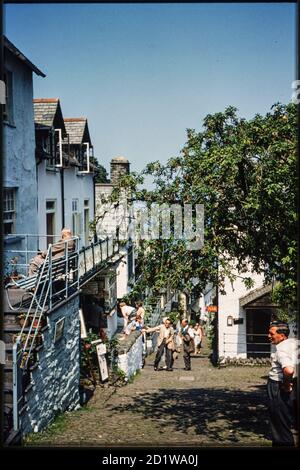 High Street, Clovelly, Torridge, Devon. Blick nach Nordosten die High Street hinunter, mit einem überhängenden Baum auf der rechten Seite und Menschen, die in der Straße gehen und sich vor einem Geschäft versammelt haben. Stockfoto