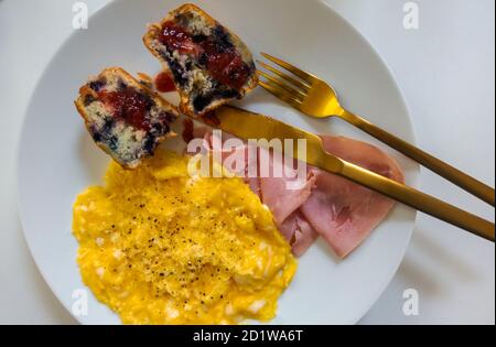 Rührei, Schinken und ein Blaubeer-Muffin auf einem weißen Teller Stockfoto