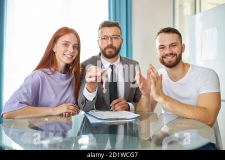 Porträt von glücklichen jungen frisch verheirateten Familie mit Immobilienmakler sitzen am Tisch zusammen, Agent in formellen Anzug halten Schlüssel in den Händen, sie lächeln an der Kamera Stockfoto