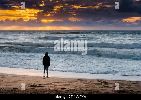 Ein Besucher, der am Fistral Beach steht und von einem spektakulären Sonnenuntergang in Newquay in Cornwall umgeben ist. Stockfoto