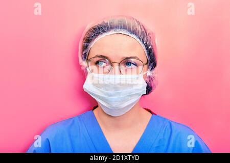 Porträt einer Ärztin mit traurigem Gesichtsausdruck, Nahaufnahme. Krankenschwester in einer blauen Uniform und einer medizinischen Maske auf einem rosa Hintergrund. Stockfoto