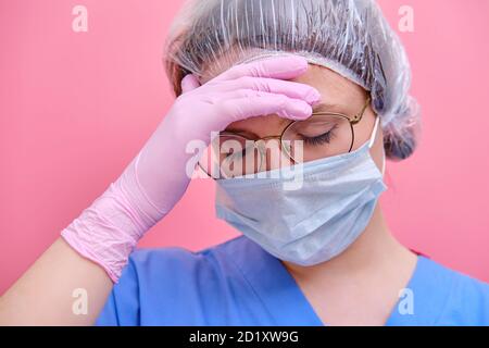 Porträt einer Ärztin mit müdem Ausdruck, Nahaufnahme. Krankenschwester in einer blauen Uniform und einer medizinischen Maske auf einem rosa Hintergrund. Stockfoto