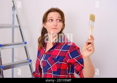 Die Malerin schaut vorsichtig auf den Pinsel gegen die weiße Wand. Probleme mit Selbstreparatur, Schwierigkeiten mit dem Anstreichen der Wände. Stockfoto