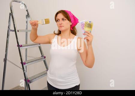 Eine Frau in einem weißen T-Shirt schaut auf eine Quaste und hält Euro-Scheine in der Hand. Vergütung von Malern und Werkstätten Stockfoto