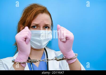 Eine Krankenschwester auf blauem Hintergrund zeigt Hände in Handschellen, Nahaufnahme. Arzt für eine Bestechung von einem Patienten verhaftet, Konzept Stockfoto