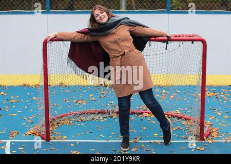 Spielplatz im Freien auf der Straße. Eine junge Frau steht am Tor des Hockeyfeldes. Herbstwetter auf dem offenen Sportplatz mit blauer Beschichtung. Stockfoto