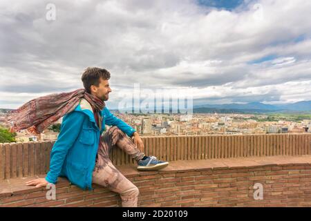 Der junge Mann mit blauer Jacke und Schal flattert im Wind und sitzt an der Wand der Festungsmauer und blickt auf das Stadtbild von Girona. Katalonien, Spanien Stockfoto