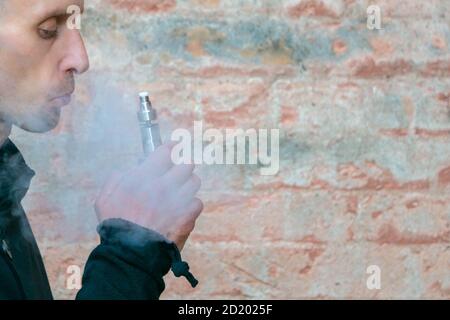 Vape man. Mann mit Dampf Mod Ausatmen Dampf der elektronischen Zigarette gegenüber Vintage-Ziegelwand. Städtischer Hintergrund mit Kopierraum. Lifestyle. Stockfoto