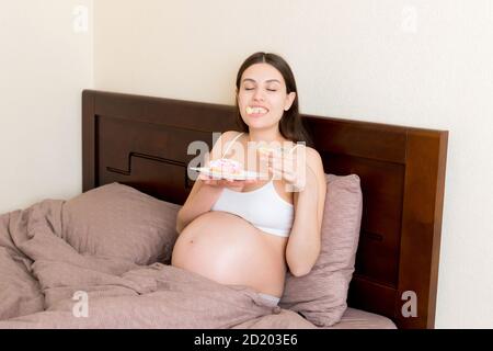 Schwangere Frau genießt essen verschiedene Donuts ruhen im Bett. Ungesunde Desserts während der Schwangerschaft Konzept. Stockfoto