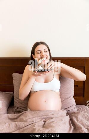 Hungrige schwangere Frau, die sich im Bett entspannt, isst gierig ein Stück Kuchen und hat einen schmutzigen Mund. Die erwartete Mutter kann nicht aufhören zu essen. Starke Appetit du Stockfoto