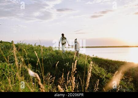 Rückansicht des jungen Mannes, der die Hand der Freundin hält, während Bei Sonnenuntergang wandern sie gemeinsam auf Hügeln Stockfoto