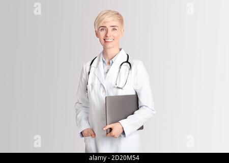 Glückliche Frau im mittleren Alter Arzt im weißen Mantel mit Stethoskop Laptop unter dem Arm halten Stockfoto