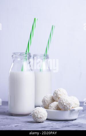 Milch in Glasflaschen mit grünen Strohhalmen mit weißer Kokosnuss Runde Süßigkeiten auf grauem Tisch auf weißem Hintergrund Stockfoto