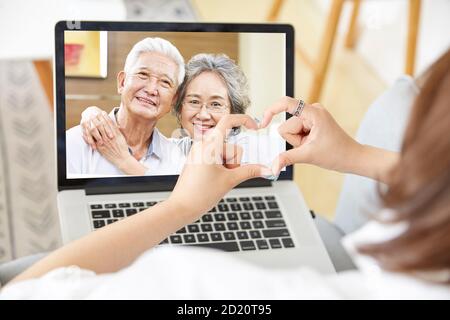 Junge asiatische Erwachsene Tochter Aufenthalt zu Hause bilden ein Herz Formen Sie sich mit den Händen, während Sie mit älteren Eltern online über sprechen Video-Chat mit Laptop-Rechner Stockfoto