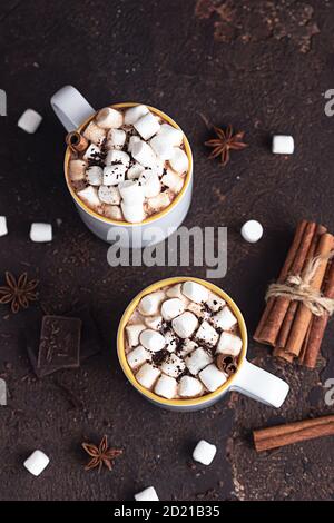 Zwei weiße Keramikbecher mit heißem Kakao oder Schokolade mit Marshmallow und Anis, Zimt und Bitterschokolade auf dunkelbraunem Steingrund. Stockfoto