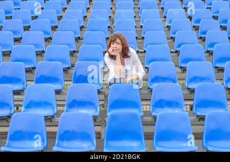 Glum Frau in leeren Zeilen des blauen Zuschauer in einem Auditorium oder Stadion mit einem gelangweilten Ausdruck und ihr Kinn auf ihren Händen sitzen Stockfoto