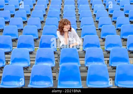 Glum Frau in leeren Zeilen des blauen Zuschauer in einem Auditorium oder Stadion mit einem gelangweilten Ausdruck und ihr Kinn auf ihren Händen sitzen Stockfoto
