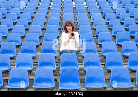 Eine Frau mittleren Alters in einem Stadion, das in der Mitte sitzt Von Reihen von leeren blauen Sitzen, die auf ihrem Handy chatten Telefon Stockfoto