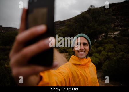 Hübscher junger männlicher Trailrunner lächelnd, der Foto von sich selbst nimmt Auf dem Smartphone auf dem Bergweg bei kaltem bewölktem Wetter Stockfoto