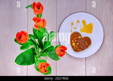 Herzförmiger Pfannkuchen und rote Blumen. Kuchen für Ihre Lieben zum Valentinstag. Ein Teller mit Pfannkuchen und Tulpen auf einem Holztisch. Ein Geschenk für den Tag o Stockfoto