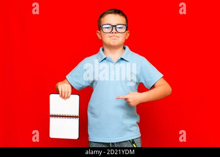 Cuccessful ernst kaukasischen Jungen in einem hellblauen T-Shirt und Brille hält, zeigt mit Finger auf leere Notebook-Banner. Boy zeigt Leerzeichen. Helle r Stockfoto