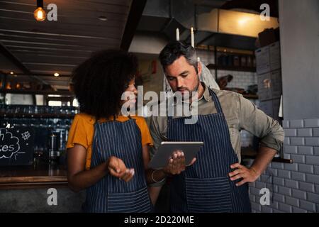 Neugierige Kollegen tragen Schürzen, diskutieren Finanzen beim Blick auf Tablet im Café stehen. Stockfoto