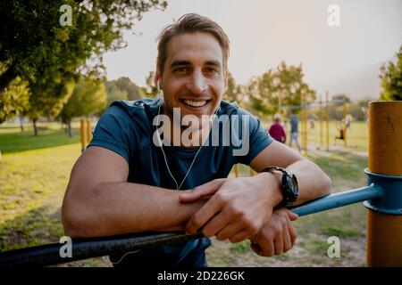 Hübscher junger männlicher Athlet lächelnd lehnt sich vor dem Freien an die Bar Workout mit Smartwatch und Ohrhörer für Musik Stockfoto