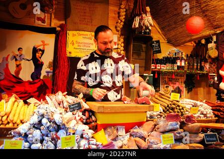 PARIS, FRANKREICH - 26. NOVEMBER 2017: Junger Metzger im lustigen T-Shirt verkauft katalanischen traditionellen Schinken und trockene Würstchen auf dem Weihnachtsmarkt in La Defense Stockfoto