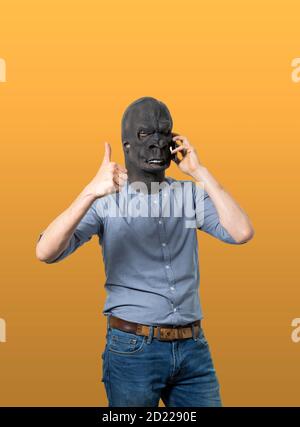 Mann in Gorilla-Maske, der am Handy spricht und den Daumen hoch gibt. Isolierter Ausschnitt auf gelbem Hintergrund. Mittlere Aufnahme. Stockfoto