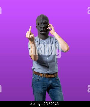 Mann in Gorilla Maske reden auf Handy geben den Mittelfinger. Isolierter Ausschnitt auf violettem Hintergrund. Mittlere Aufnahme. Stockfoto