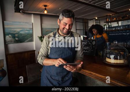 Schöner und lächelnder männlicher Kaffeehaus-Besitzer, der im Café auf dem Tablet browst. Stockfoto