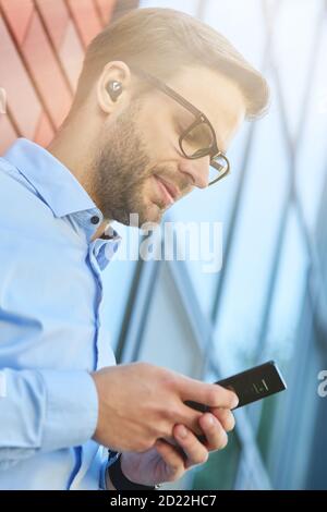 Seitenansicht eines jungen, hübschen Geschäftsmannes, der schnurlose Ohrhörer trägt Mit seinem Smartphone, während er sich im Freien an einer Ziegelwand lehnt Stockfoto