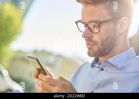 Seitenansicht eines jungen gutaussehenden Geschäftsmann mit Smartphone während Sitzen auf der Bank im Freien an einem sonnigen Tag Stockfoto