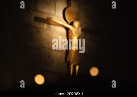 Verschwommenes Foto des Kruzifixes an der Steinwand und der Kerzen im Vordergrund. Symbolischer Kontrast von Licht und Dunkelheit. Stockfoto