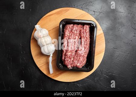 Hackfleisch in einem Kunststofftablett und ganze weiße Knoblauchköpfe in einem Netzbeutel auf Holz Schneidebrett auf schwarzem Stein Hintergrund, Draufsicht. Frisch, roh, gehackt Stockfoto