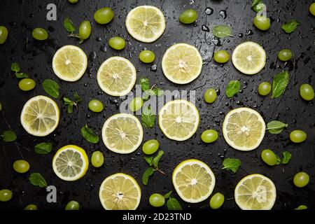 Zitronenscheiben, grüne Minzblätter und Trauben auf schwarzem Hintergrund mit Wassertropfen, Draufsicht. Frische tropische Früchte, gelbe Zitrusfrüchte und Beeren, flaches Lay Stockfoto